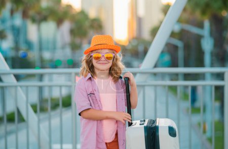 Foto de Divertido viajero infantil. Niño turista viajero en ropa casual, gafas de sol de moda y sombrero sostienen maleta. Viaja al extranjero los fines de semana. Viaje de niños - Imagen libre de derechos