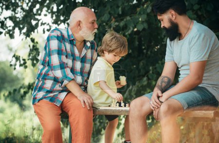 Handsome grandpa and grandson are playing chess while spending time together outdoor. Boy with father and grandfather. Father and son in the park