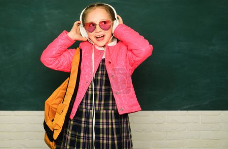 Photo for Teenager younf school girl with backpack on blackboard. Portrait of a teen female student. Funny school girl wearing eyeglasses. Young teenager schoolgirl with school backpack and headphone - Royalty Free Image