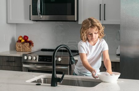 Téléchargez les photos : Nettoyage enfant vaisselle avec éponge. Du matériel de nettoyage. Aide à nettoyer. Tâches ménagères. Il lave la vaisselle. Aide aux enfants pour laver la vaisselle à la cuisine - en image libre de droit