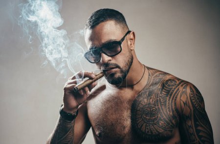 Téléchargez les photos : Portrait d'un bel homme fumant du cigare. Homme tatoué avec cigare. Portrait d'un patron de la mafia avec un torse nu fort fumant un cigare. Chef de la mafia - en image libre de droit
