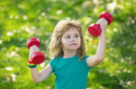 Photo for Child boy raising a dumbbell. Cute child training with dumbbells. Children fitness. Kid boy exercising with dumbbells outdoor. Funny excited child sportsman - Royalty Free Image