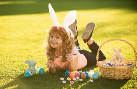 Foto de Niño niño con huevos de Pascua y orejas de conejo que ponen en la hierba pintando huevos - Imagen libre de derechos