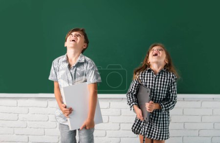 Foto de Los niños de la escuela tienen un libro con una expresión sorprendente contra la pizarra. Escuela niños amigos - Imagen libre de derechos