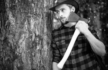 Woodcutter with axe in the summer forest. Lumberjack standing with axe on forest background. Lumberjack in the woods with an ax