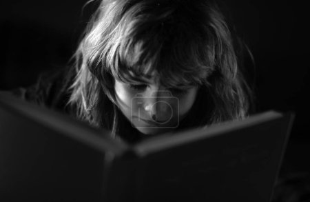 Téléchargez les photos : Enfant lisant l'histoire du coucher dans la chambre noire. Un petit garçon mignon lisant un livre allongé sur le lit. Enfant lisant un livre le soir du coucher - en image libre de droit