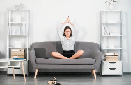 Young attractive secretary woman doing yoga exercise on sofa at workplace in a modern office. Pretty accountant girl meditating on office. Employee feel balance harmony relaxation