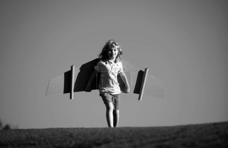 Los niños vuelan. Divertido niño piloto volando con alas de avión de cartón de juguete en el cielo azul, espacio de copia. Inicie el concepto de libertad, niño despreocupado