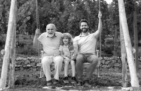 Photo for Men generation family playing on swing together Three different generations ages: grandfather father and son swinging together in summer garden outdoors. Active summer leisure for family - Royalty Free Image