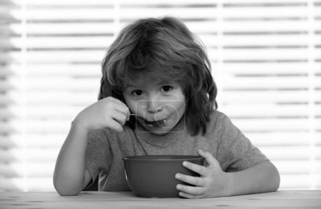 Téléchargez les photos : Portrait d'enfant mignon mangeant un repas de soupe ou petit déjeuner déjeuner à la table à la maison avec cuillère. Enfants alimentation saine - en image libre de droit
