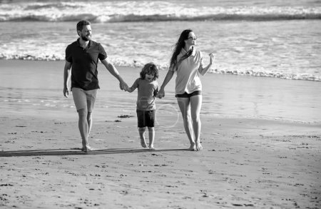 Foto de Longitud total de la familia de tres caminando a lo largo de la orilla del mar. Padres tomados de la mano del hijo y caminando en la playa - Imagen libre de derechos