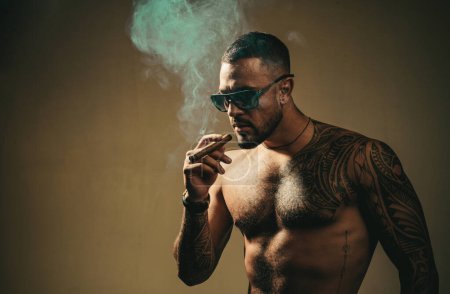 Tätowierter Mann mit Zigarre. Zigarrenraucher genießen das Leben und den Moment. Porträt eines bärtigen Geschäftsmannes mit schönem Oberkörper, der eine Zigarre raucht und Whiskey trinkt.