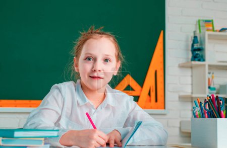 Foto de Little girl pupil with happy face expression near desk with school supplies. Learning concept. Happy school kids at lesson - Imagen libre de derechos