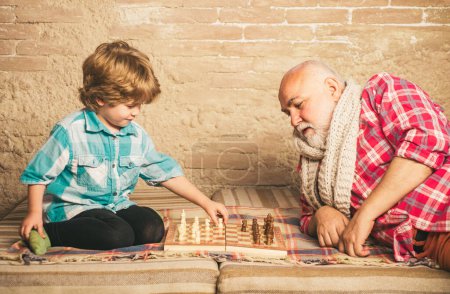 Téléchargez les photos : Senior enseignant à son petit-fils à jouer aux échecs. Beau grand-père et petit-fils jouent aux échecs tout en passant du temps ensemble à la maison - en image libre de droit