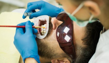 Téléchargez les photos : Dentiste examinant les dents des patients à la clinique. Homme barbu ayant des dents examinées chez les dentistes. Jeune homme souriant après la visite dentiste sur fond flou lumineux - en image libre de droit