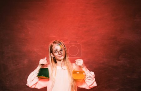 Photo for Pretty little girl learning chemistry isolated at red background. Young scientist girl in protective glasses making experiment in lab or chemical cabinet. Child holding two big tubes - Royalty Free Image