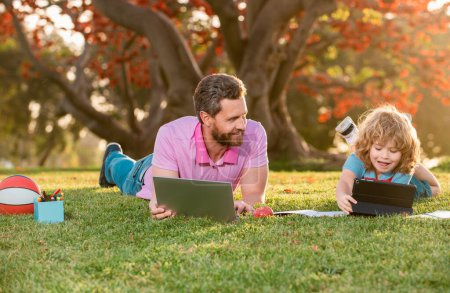 Foto de Niño feliz con el padre aprendiendo al aire libre estudiando en línea y trabajando en el ordenador portátil en el campo verde - Imagen libre de derechos