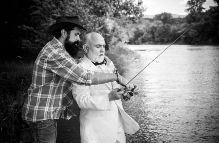 Téléchargez les photos : Père et fils mature pêcheur pêche avec une canne à pêche sur la rivière. Portrait de l'homme d'affaires senior pêche en costume blanc et noeud papillon avec canne à pêche, bobine filante sur la rivière - en image libre de droit