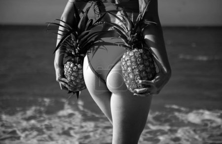 Foto de Sexy woman in swimsuits bikini hold pineapple near butt, buttocks on summer beach - Imagen libre de derechos