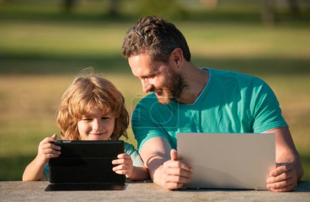 Téléchargez les photos : Père et fils jouent ou étudient avec un ordinateur portable dans le parc - en image libre de droit