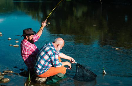 Téléchargez les photos : Heureux grand-père et petit-fils avec des cannes à pêche sur le quai de la rivière. Hommes pêchant dans la rivière pendant la journée d'été. Père et fils se relaxent ensemble - en image libre de droit