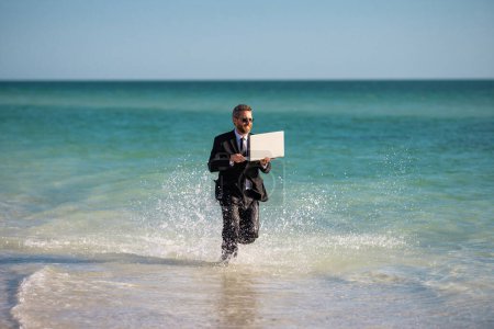 Photo for Businessman tourist in casual suit barefoot walking on tropical beach with laptop. Handsome business man on summer vacation. Businessman running on sea holding laptop. Businessman jumping in sea - Royalty Free Image