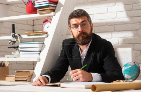 Photo for Portrait of thinking male teacher in school classroom. Serious teacher man sitting on desk writing text in book at school classroom - Royalty Free Image
