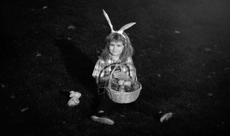 Foto de Niño con huevos de Pascua en cesta al aire libre. Niño en el césped en el parque. Caza de huevos de Pascua. Retrato de niños Fynny. Lindo conejito niño con orejas de conejo - Imagen libre de derechos