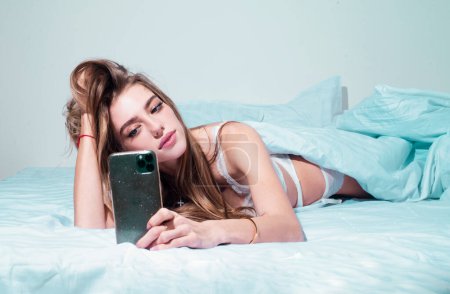 Foto de Chica joven charlando por teléfono por la mañana en la cama. Mujer en la cama revisando aplicaciones sociales con smartphone - Imagen libre de derechos