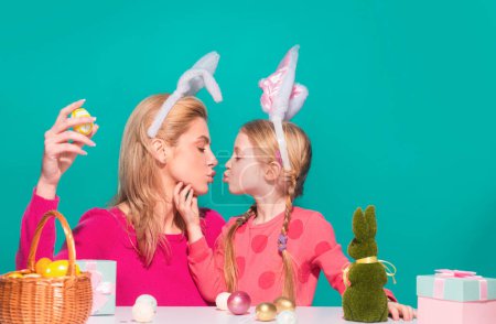 Foto de Feliz Pascua. Una madre y su hija pintando huevos de Pascua y besándose. Familia feliz preparándose para la Pascua. Linda niña pequeña con orejas de conejo en el día de Pascua aislado en azul - Imagen libre de derechos