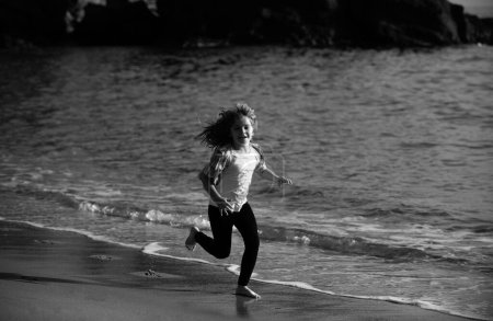 Foto de Niño feliz corriendo en la playa del mar. Chico gracioso corre a lo largo del borde del surf. Estilo de vida de niños activos. Carrera y deporte saludable para los niños - Imagen libre de derechos