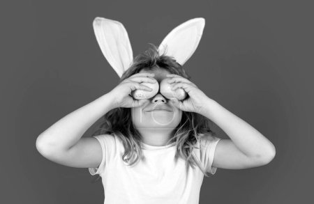 Kids boy in bunny ears hold near eyes easter eggs. Funny kids face