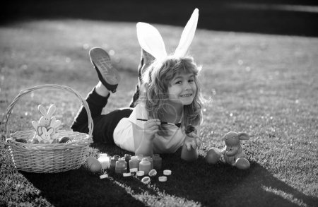 Téléchargez les photos : Enfant de Pâques garçon dans des oreilles de lapin peignant des œufs de Pâques en plein air. Enfant mignon en costume de lapin avec des oreilles de lapin s'amusant dans le parc - en image libre de droit