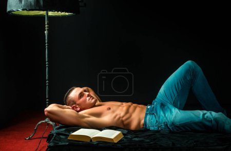 Foto de Hombre guapo en denim azul con libro y torso desnudo. Acuéstate en la cama y relájate. Estilo de vida de lujo - Imagen libre de derechos