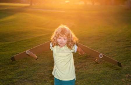 Téléchargez les photos : Enfant jouant avec jetpack jouet au champ d'herbe coucher de soleil. Enfant pilote astronaute ou astronaute rêve de vol. Aviateur garçon volant avec des ailes en carton - en image libre de droit