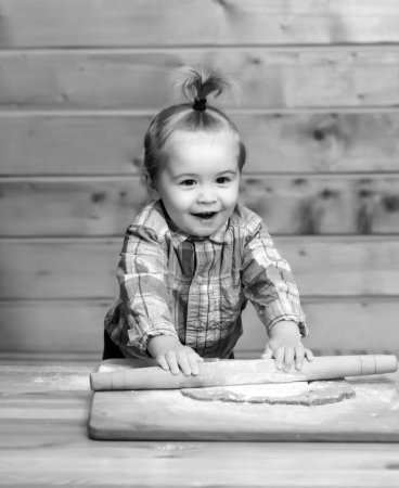 Téléchargez les photos : Image amusante de bébé chef jouer avec de la farine sur fond de cuisine en bois. Portrait de jeune enfant faisant de la boulangerie maison dans la cuisine. Enfant assis sur la table et profiter d'apprendre à cuisiner des aliments ou de cuisson - en image libre de droit