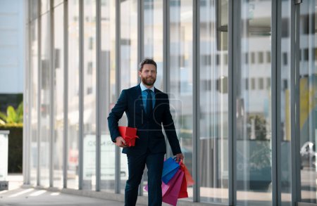 Photo for Business man shopping in a shopping center. Happy businessman in suit holding paperbags. Shopaholic man walking on commercial street with a lot of shopping bags. Rich man - Royalty Free Image