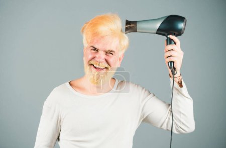 Foto de Elegante hombre con secador de pelo y expresiones divertidas en la barbería. Rubia barbudo hombre cabello seco - Imagen libre de derechos