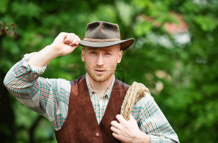Téléchargez les photos : Cowboy farmer man in country side wearing western cowboy hat. Cowboy with lasso rope on green background. American Male model in countryside on farm - en image libre de droit