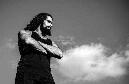 Young muscular handsome man standing with arms folded against blue sky looking forward copy space and space for text. Fashionable man with long wavy hair and strong healthy body