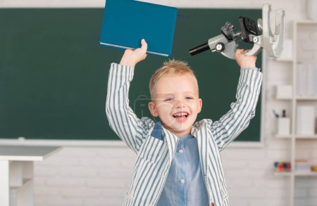 Photo for Little schoolboy study in a classroom at elementary school. Excited schoolboy pupil with microscope and book - Royalty Free Image