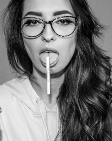 Foto de Atractiva chica hipster en gafas sosteniendo cigarrillo en la lengua. Hermosa morena sexy con un elegante retrato de moda cigarrillo. Mujer con adicción a la nicotina - Imagen libre de derechos