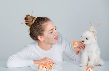 Foto de Belleza mujer sexy con perro comiendo sushi. Rollos de sushi. Chica modelo de moda comiendo Sushi con palillos. Maquillaje perfecto y corte de pelo bob con palillos, comida japonesa - Imagen libre de derechos