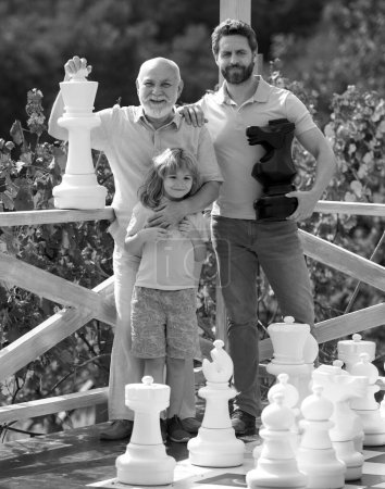 Foto de Abuelo con hijo y nieto jugando ajedrez. Hombres asombrados en diferentes edades jugando ajedrez juego de mesa. Tres generaciones de hombres. Concepto del día de los padres - Imagen libre de derechos