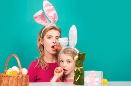 Loving family concept. Happy mother with her daughter with bunny ears are preparing for Easter. Funny bunny girls eating carrot