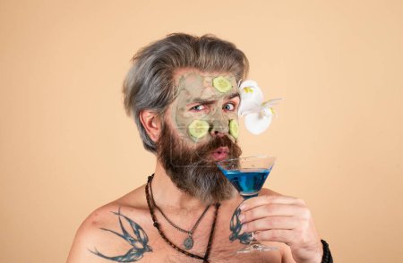 Lustiger Mann mit Lehmmaske und Gurkenscheiben im Gesicht trinkt Cocktail. Spa, Dermatologie, Wellness und Gesichtsbehandlung. Mann mit kosmetischer Feuchtigkeitsmaske, isolierter Studiohintergrund