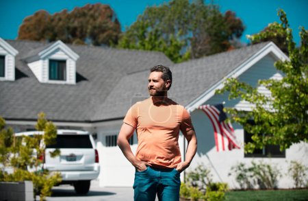 Foto de Retrato del tipo risueño con las manos en los bolsillos apoyadas en la casa americana. Un hombre parado frente a su nuevo hogar. Gente real - Imagen libre de derechos