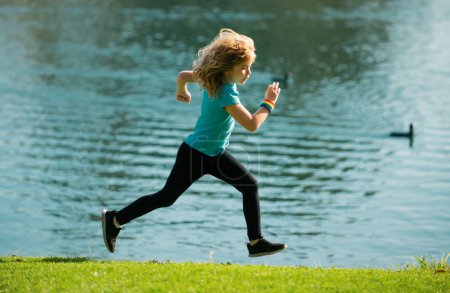 Téléchargez les photos : Enfants courir ou faire du jogging près du lac sur l'herbe dans le parc. Garçons jogging coureur dans le parc extérieur. La course est un sport qui renforce le corps. Sports de plein air et fitness pour les enfants, exercice à l'extérieur - en image libre de droit