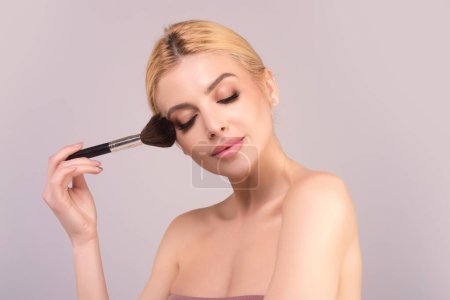 Studio portrait of a woman applying cosmetic tonal foundation on face using makeup brush. Beautiful girl doing contouring apply blush on cheeks isolated on studio background