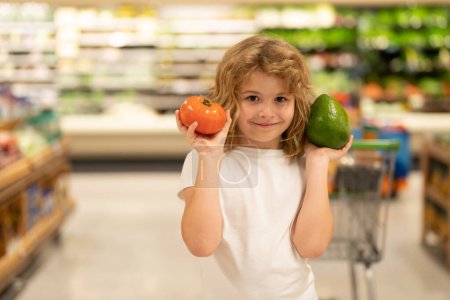 Téléchargez les photos : Enfant choisissant des légumes en magasin alimentaire. Des enfants au marché alimentaire. Ventes, rabais dans le magasin du marché alimentaire et les achats. Garçon mignon dans le marché détiennent des légumes et des fruits. Shopping d'enfants dans le magasin du marché - en image libre de droit